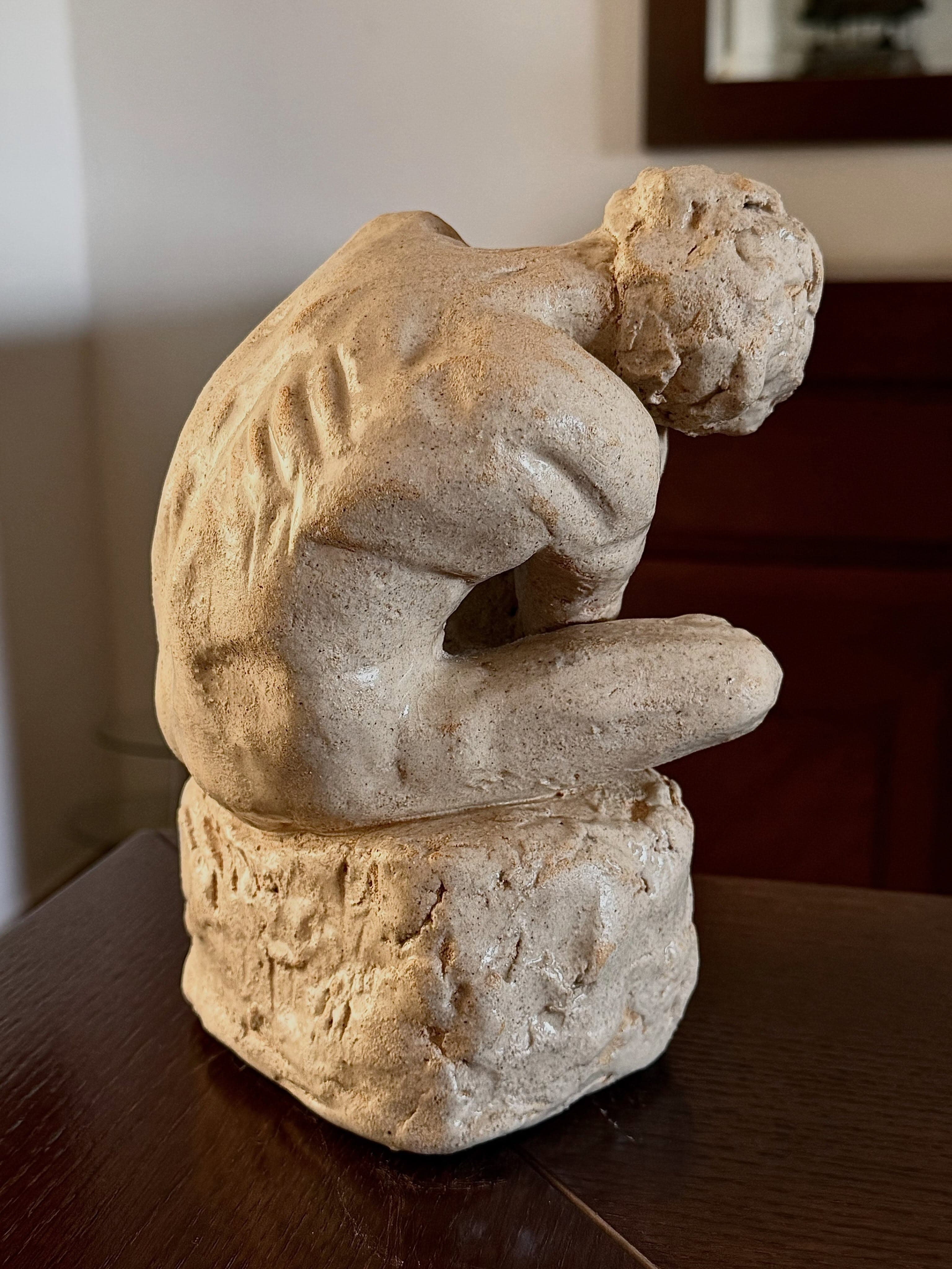 A figure crouched and curled into itself in pale clay, head bowed onto folded arms and drawn-up knees, seated on a rough rock block. Deep parallel ridges are scored down the curve of its back.